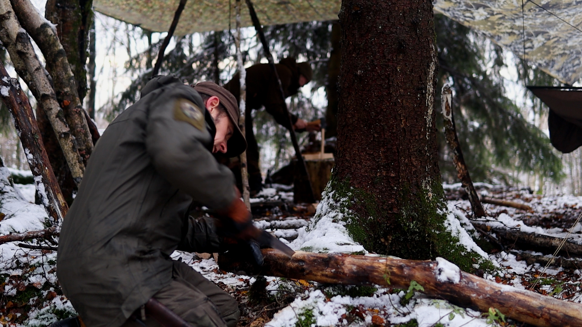 Person hackt Brennholz unter dem aufgespannten Tarp im verschneiten Winterwald