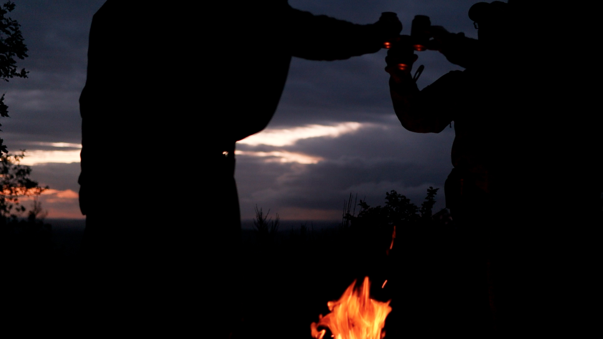 Drei Bushcrafter stoßen mit Bierflaschen an — Silhouette gegen den Abendhimmel in der Hocheifel
