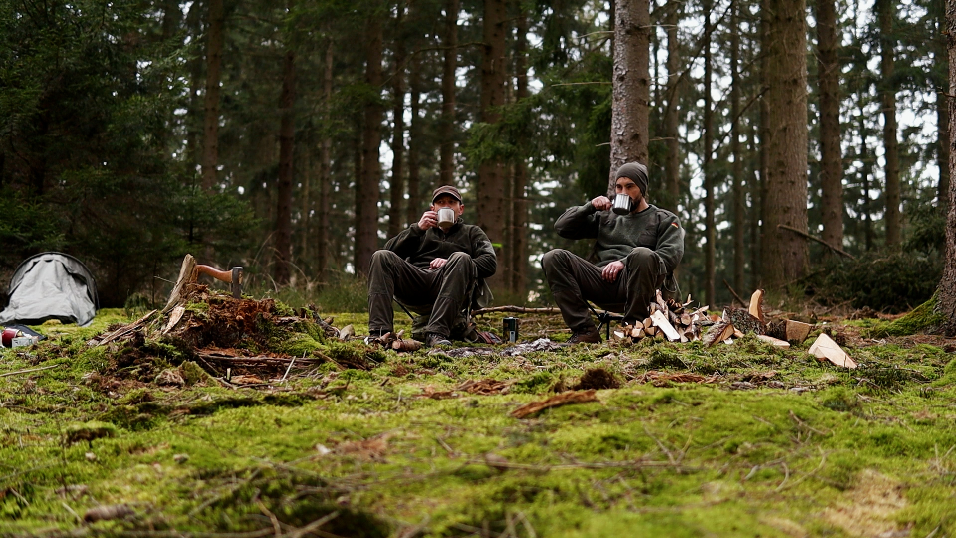 Zwei Bushcrafter trinken aus Bechern im moosigen Nadelwald
