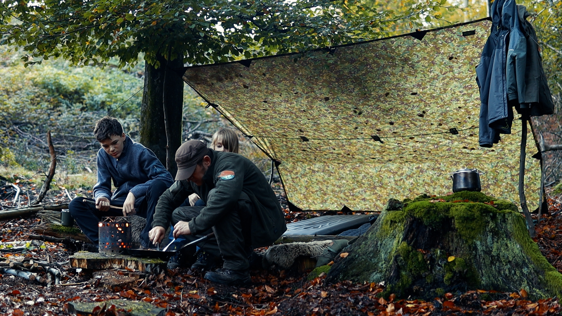 Vater und Söhne entzünden das Feuer unter dem aufgespannten Tarp