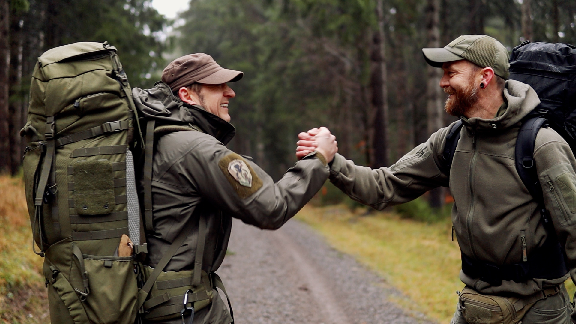 Zwei Bushcrafter mit Rucksäcken geben sich die Hand auf einem Waldweg im Regen