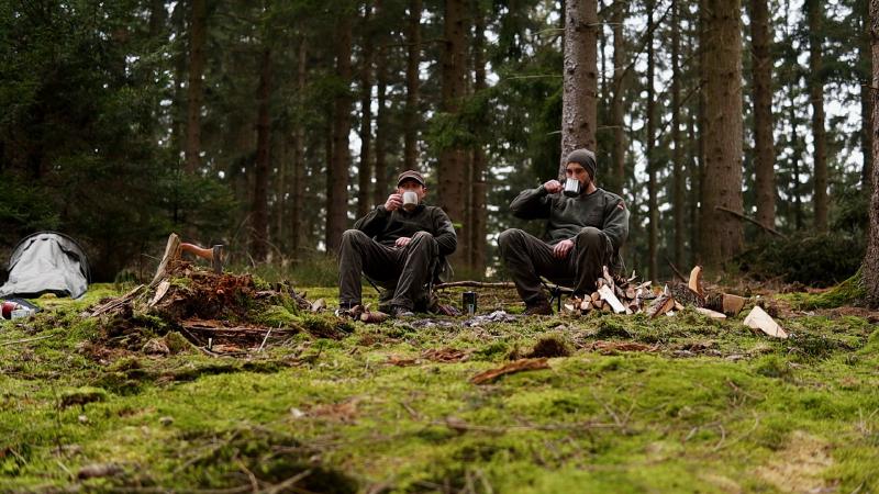 Zwei Bushcrafter trinken aus Bechern auf dem moosigen Waldboden